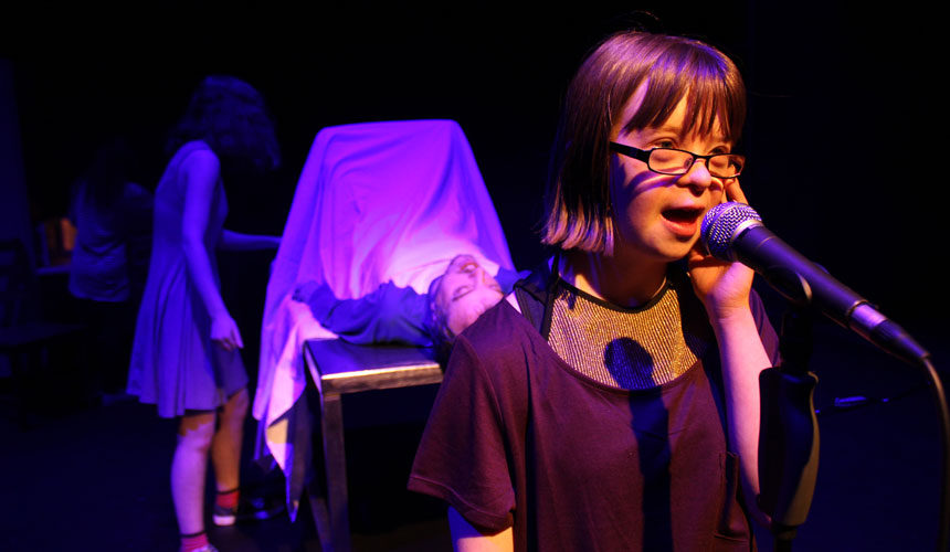 A woman speaks into a microphone. A man is on a table behind her with a white sheet covering him from the chest down.