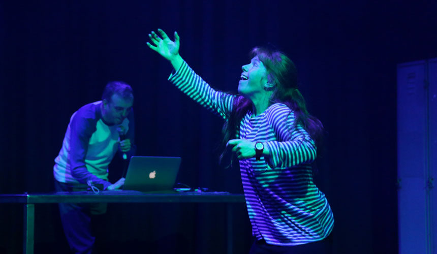 A woman dancing and reaching upwards. A man is in the background on a computer.